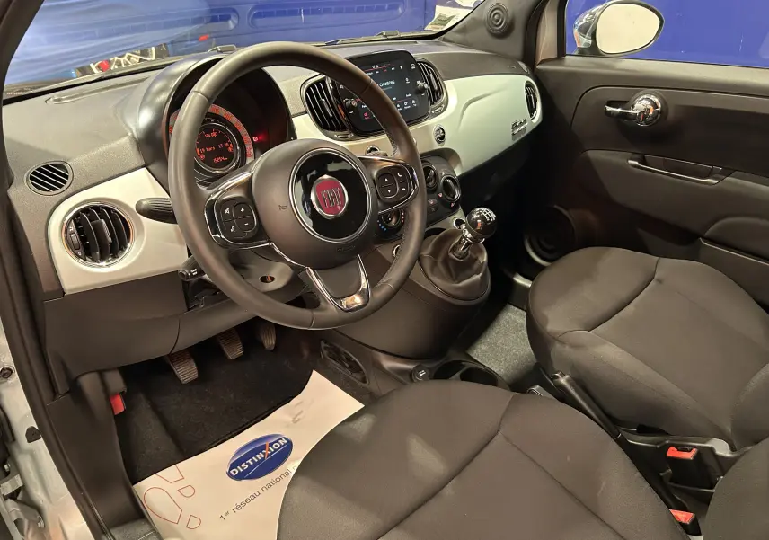 Vue intérieure côté conducteur de la Fiat 500 2023, tableau de bord clair avec volant multifonction et levier de vitesse manuel.