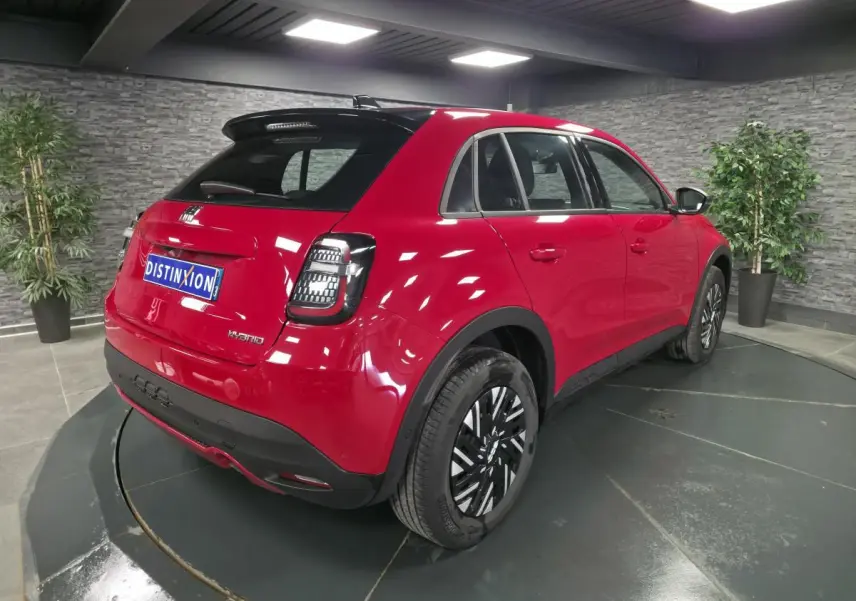 Vue 3/4 arrière droite d'une FIAT 600 rouge hybride 2024 avec jantes bicolores et toit noir brillant en showroom.