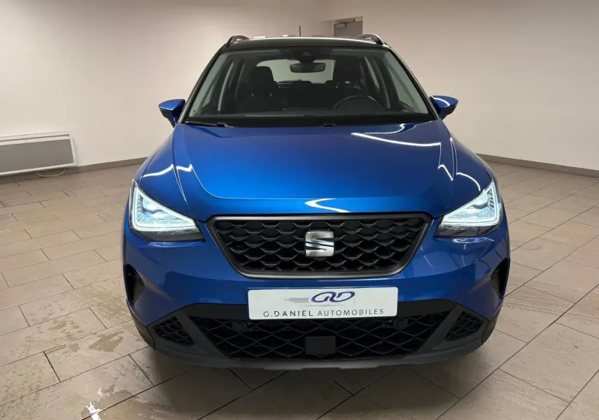 Vue avant d’un SEAT Arona bleu saphir avec toit noir, phares allumés et calandre noire en intérieur neutre.