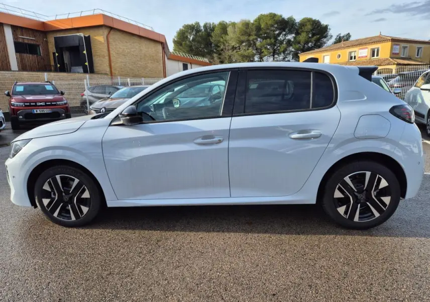 Profil droit d'une Peugeot 208 Hybrid 110 Allure blanche avec jantes alliage 16 pouces, vue en extérieur sur parking.