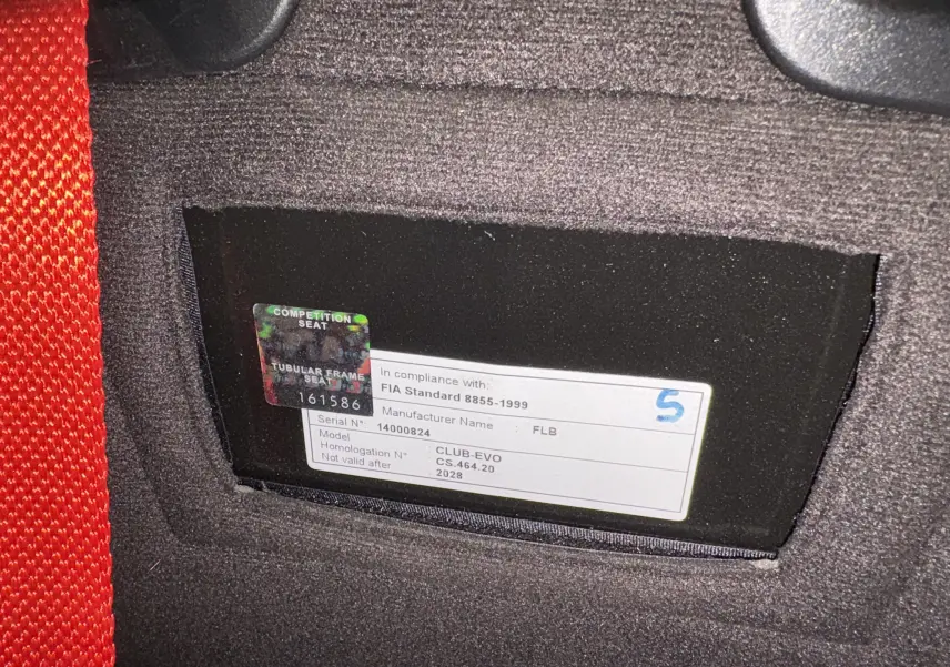 Gros plan sur l’étiquette FIA d’un siège baquet rouge de compétition dans un Audi Coupé GT noir de 1983.