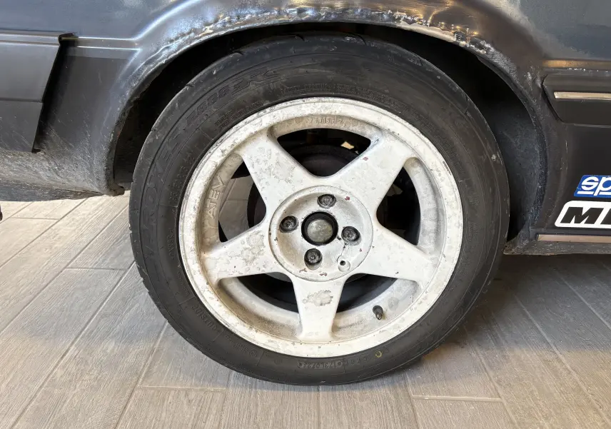 Gros plan sur la roue avant gauche d’un Audi Coupé GT noir de 1983 avec jante blanche cinq branches usée et carrosserie marquée.