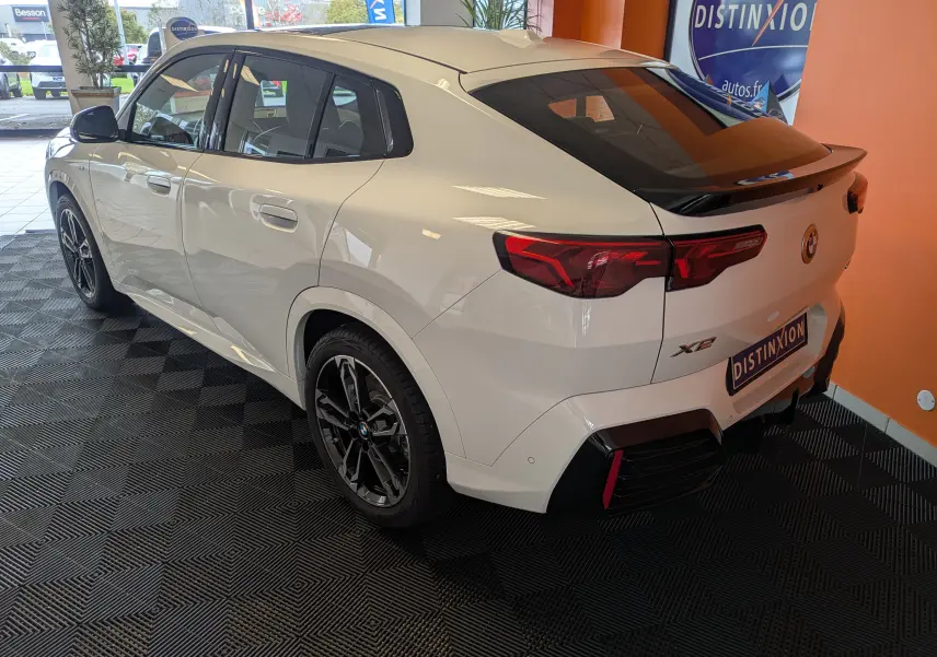 BMW X2 sDrive blanc alpin vu de l'arrière côté gauche, avec jantes noires et toit panoramique électrique visible.