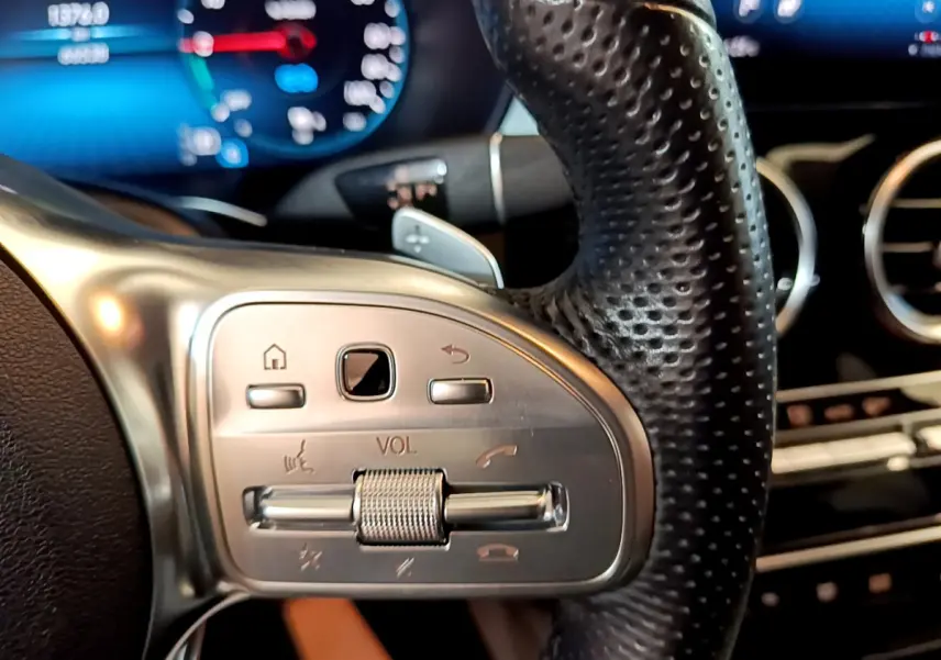 Gros plan sur les commandes du volant en cuir perforé du Mercedes GLC 300 E AMG Line 2021, avec tableau de bord numérique flou.