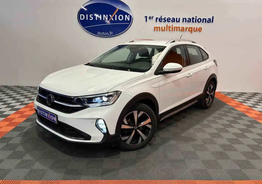 Volkswagen Taigo TSI 110 blanc pur vu en 3/4 avant droit avec jantes noires et phares allumés dans un showroom.