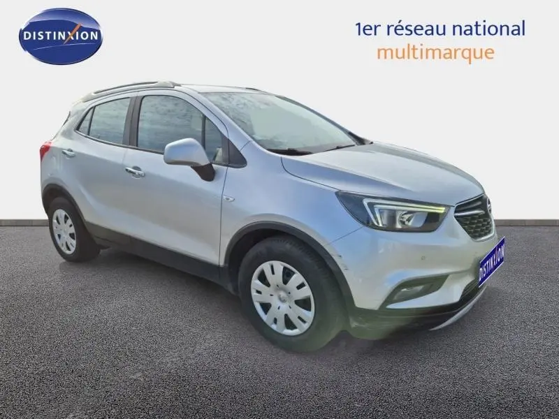 Opel Mokka X gris minéral en 3/4 avant droit, avec barres de toit et jantes acier, sur fond neutre.