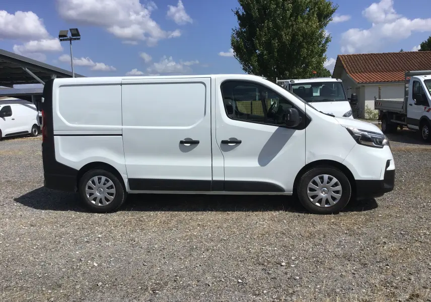Profil droit d'un utilitaire blanc Nissan Primastar Fourgon L1H1 2022, stationné sur un sol gravillonné par temps ensoleillé.