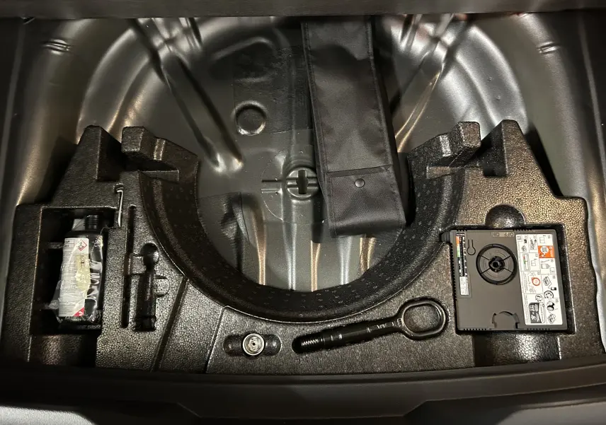 Vue plongeante sur le coffre ouvert du Volkswagen T-Roc gris Indium, montrant le rangement pour roue de secours et outils.