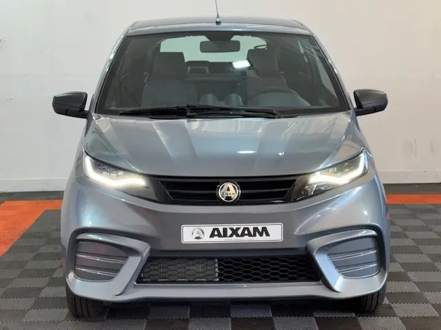Vue frontale d'une Aixam Ambition 0.5 City Pack gris foncé 2026 avec phares allumés et rétroviseur caméra côté gauche.