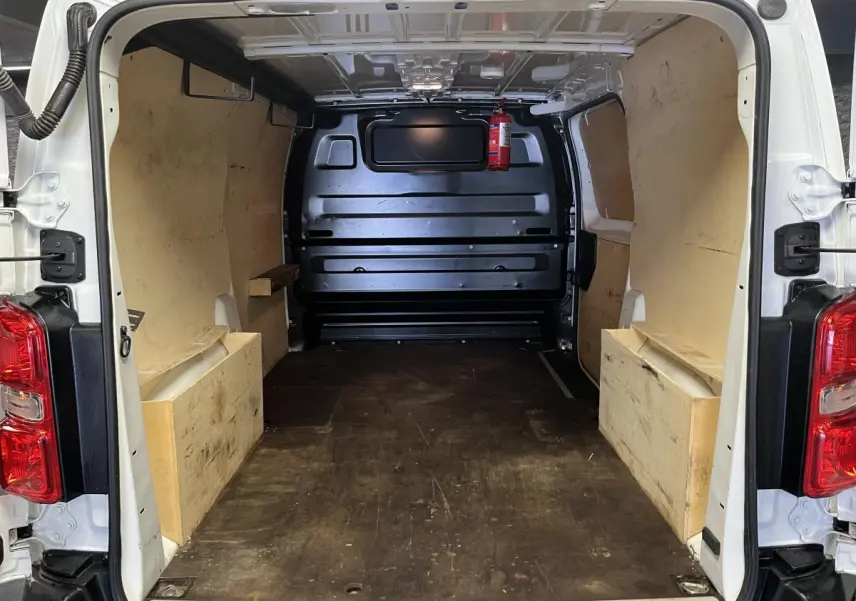 Intérieur du coffre arrière ouvert d'un Citroën Jumpy blanc, montrant le plancher et habillage bois latéral.