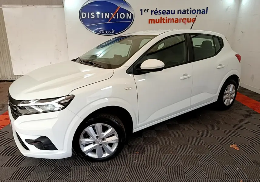 Vue 3/4 avant d'une Dacia Sandero Confort TCe 90 blanche, stationnée en intérieur sur sol damier noir et rouge.