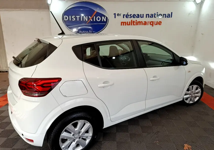 Vue 3/4 arrière droite d'une Dacia Sandero Confort blanche 2022 dans un showroom avec logo Distinxion en arrière-plan.