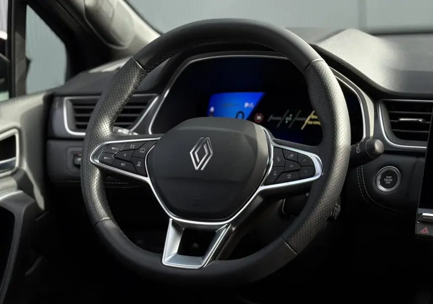 Vue rapprochée du volant noir perforé avec logo Renault et tableau de bord numérique bleu en arrière-plan.