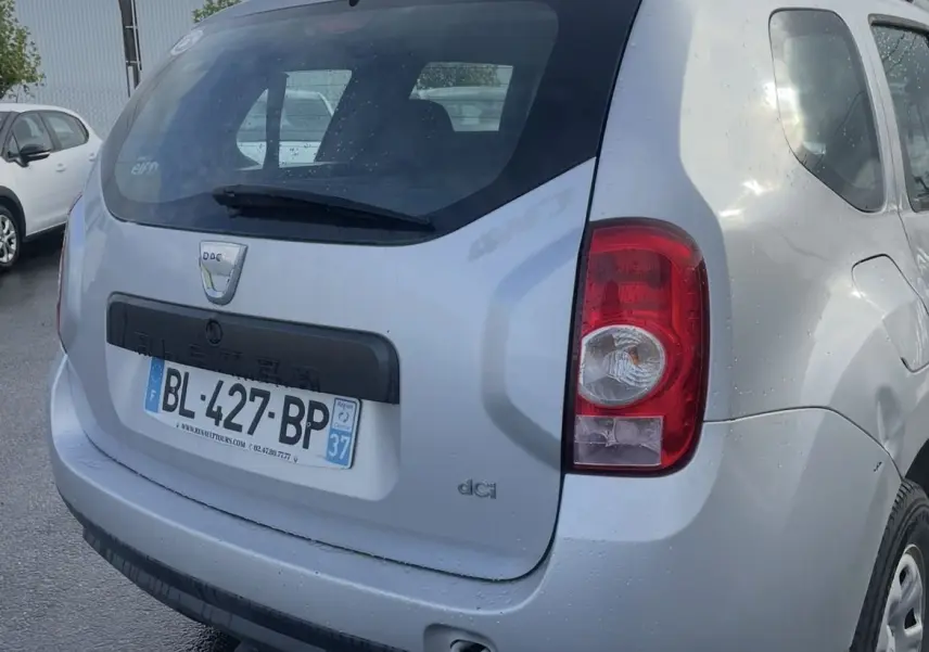 Vue arrière droite d'un Dacia Duster gris 2011 avec attelage et barres de toit sous un ciel nuageux.