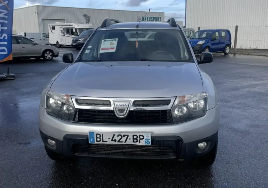 Vue frontale d'un Dacia Duster gris 2011 avec barres de toit et plaque d'immatriculation française visible.