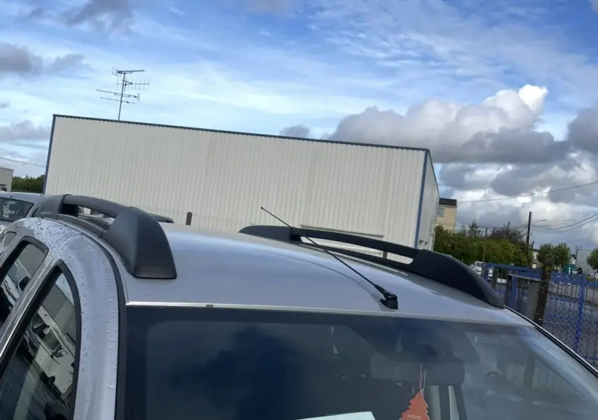 Vue 3/4 avant droit d'un Dacia Duster gris 2011 avec barres de toit et étiquette de vente visible sur le pare-brise.