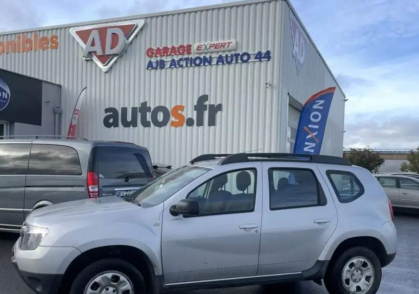 Profil côté gauche d'un Dacia Duster gris 2011 avec barres de toit et enjoliveurs, stationné devant un garage autos.fr.