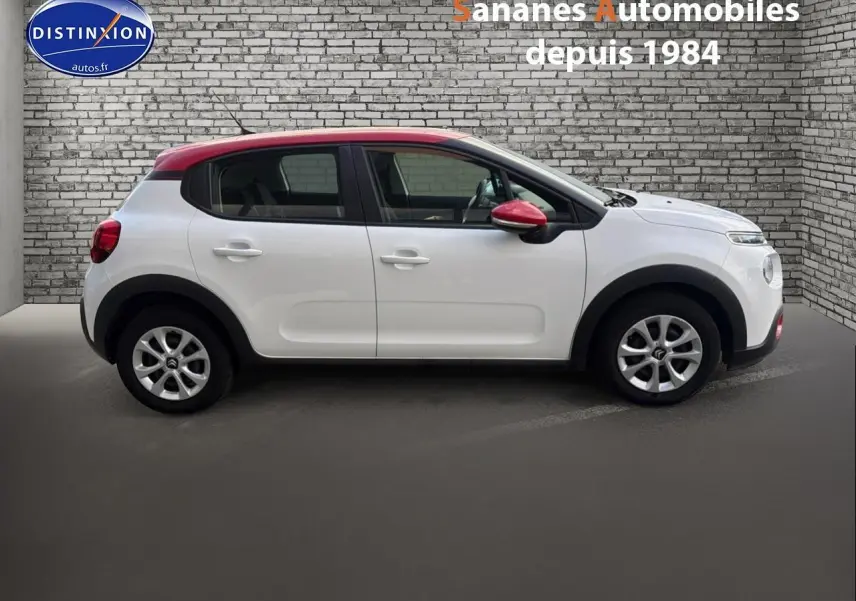 Profil droit d'une Citroën C3 blanche avec toit et rétroviseur rouge, sur fond de mur en briques grises.