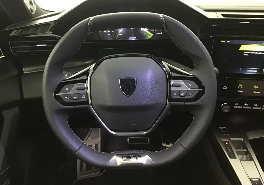 Volant cuir noir GT de la Peugeot 308 2025 avec commandes intégrées et tableau de bord numérique en arrière-plan.