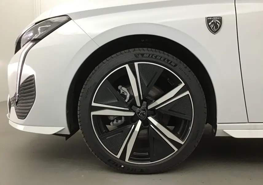 Gros plan sur la roue avant gauche d'une Peugeot 308 blanche Okenite avec jante alliage noire et argentée et logo lion.