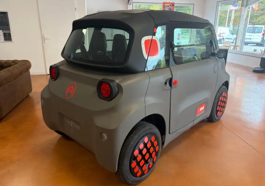 Vue 3/4 arrière droite de la Citroën Ami Spicy 2025 gris mat avec détails rouges sur roues et carrosserie.