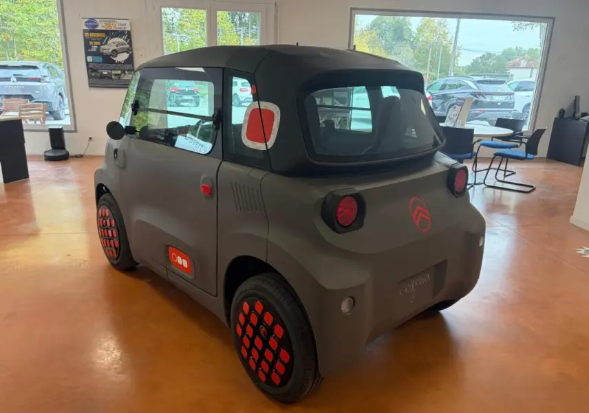Vue 3/4 arrière droite de la Citroën AMI Spicy 2025 gris avec détails rouges distinctifs et toit noir en intérieur showroom.