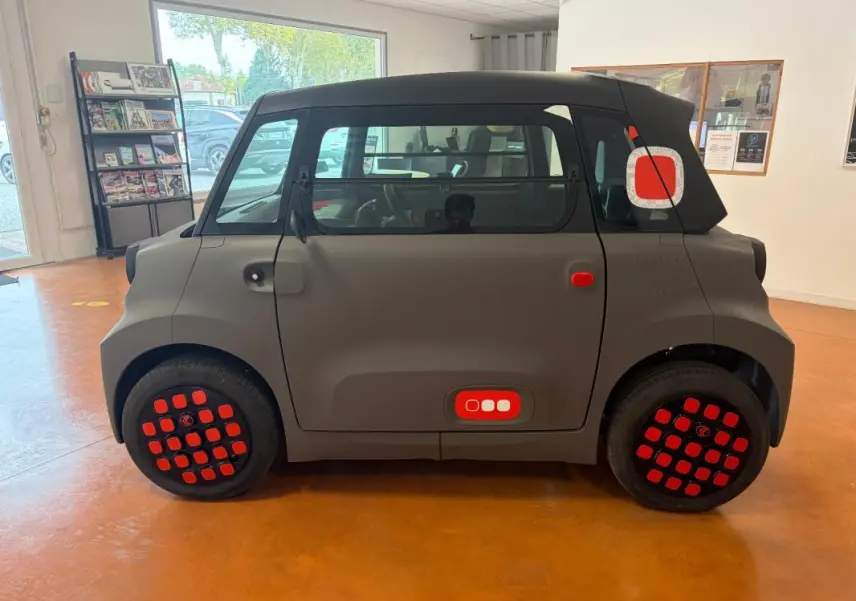Vue de profil côté gauche d'une Citroën Ami Spicy 2025 gris mat avec détails rouges sur les roues et la porte arrière.