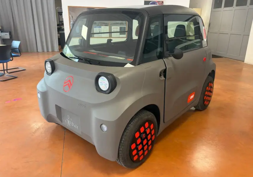 Vue 3/4 avant droite du Citroën Ami Spicy gris avec détails rouges sur jantes et logo, dans un intérieur lumineux.