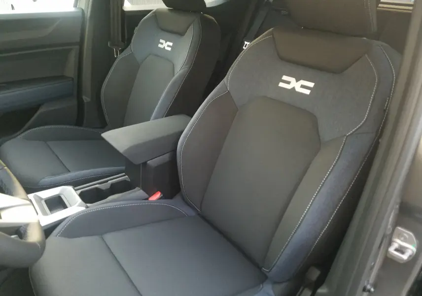 Gros plan sur les sièges avant gris foncé du Dacia Duster 2025 avec logo DC brodé et accoudoir central.