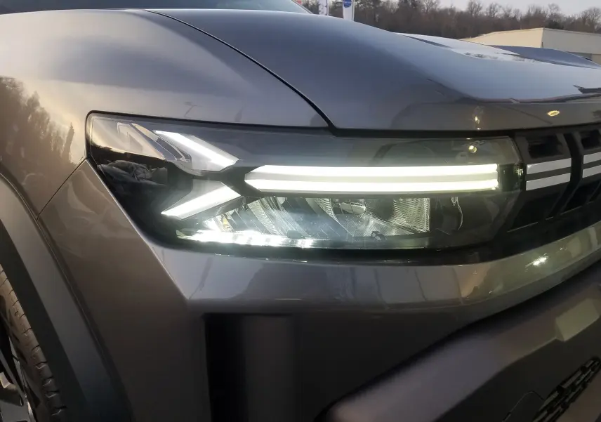 Gros plan sur le phare avant droit allumé du Dacia Duster 2025 gris schiste avec signature lumineuse LED distinctive.