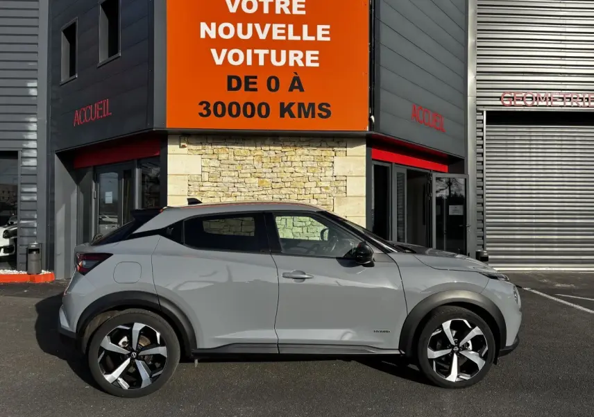 Profil droit d'un Nissan Juke Hybrid gris clair 2024 avec jantes alliage noires et toit noir, stationné devant un bâtiment.