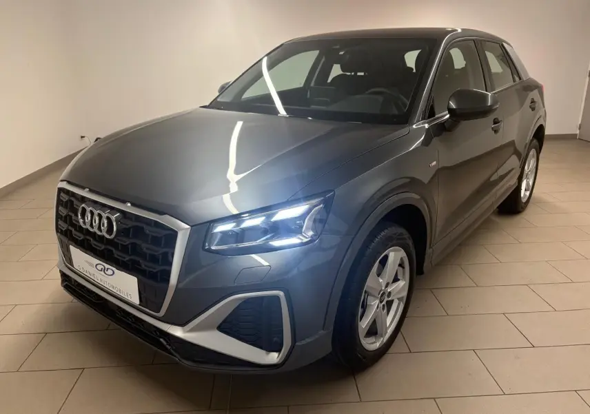 Audi Q2 35 TFSI S Line gris Daytona en 3/4 avant droit, phares allumés et calandre noire distinctive.