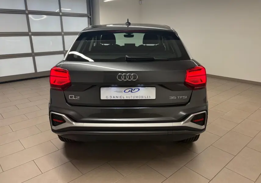Vue arrière d'un Audi Q2 35 TFSI S Line gris Daytona métallisé, avec feux rouges et pare-chocs argenté en intérieur showroom.