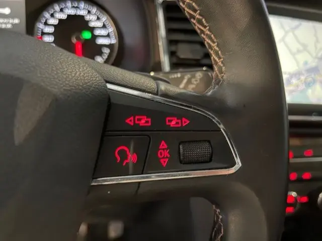 Gros plan sur les commandes rouges du volant en cuir noir du SEAT Ateca blanc Nevada, avec tableau de bord et écran GPS en arrière-plan.