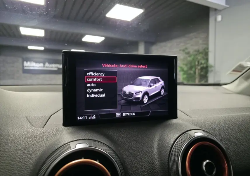 Écran MMI affichant Audi Q2 blanc en vue 3/4 avant, avec tableau de bord noir et aérateurs cerclés de rouge.