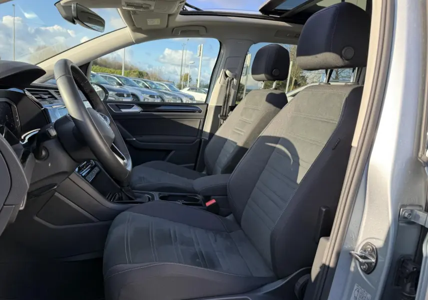 Vue intérieure côté conducteur du Volkswagen Touran argent Dolomite 2025 avec toit ouvrant panoramique et sellerie microfleece noire.