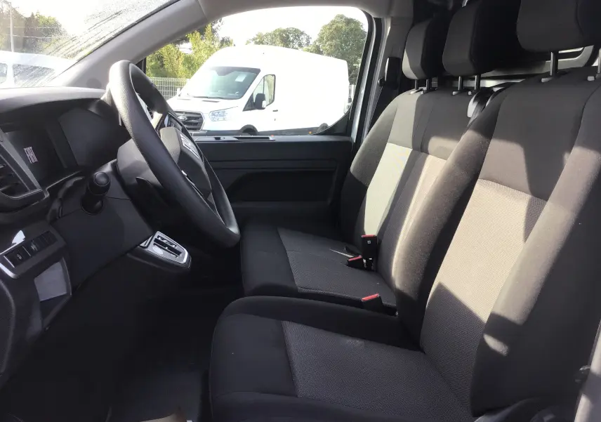 Vue intérieure côté conducteur du Fiat Scudo Fourgon blanc 2024, montrant les sièges avant en tissu noir et le volant.