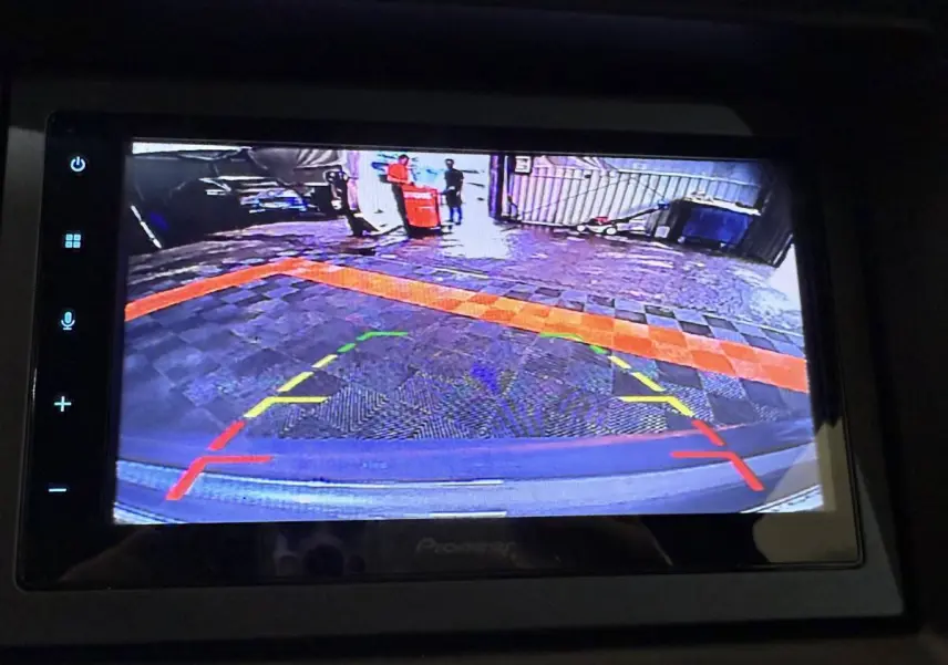 Vue écran caméra de recul côté conducteur d'une Aixam Ambition 0.5 Crossover Premium gris foncé, affichant le sol pavé et une barrière.