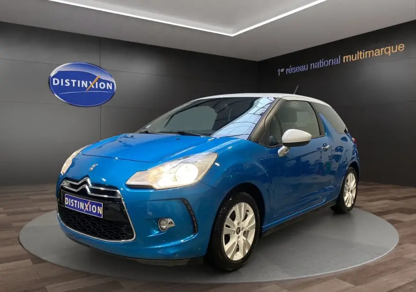 Citroën DS3 bleu avec toit blanc en 3/4 avant droit, phare allumé et jantes blanches visibles en showroom.