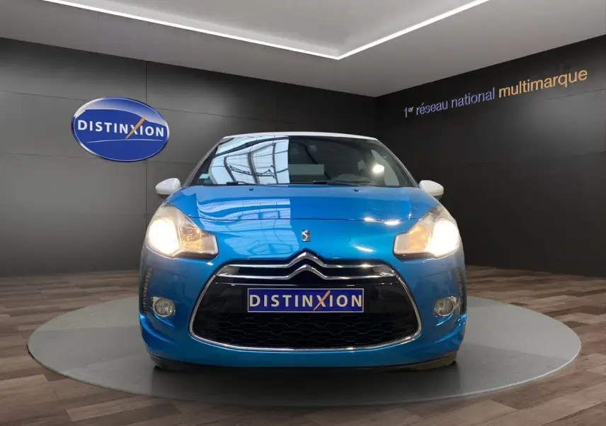 Vue avant d'une Citroën DS3 bleu avec toit blanc, phares allumés, sur fond intérieur showroom Distinxion.