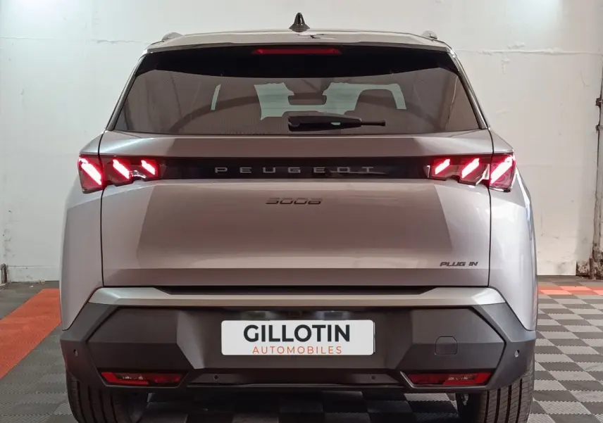 Vue arrière d’un Peugeot 5008 gris clair 2026 avec feux LED allumés et logo PLUG IN visible à droite.