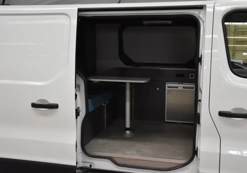 Vue latérale droite du Renault Trafic Combi blanc 2021 avec porte coulissante ouverte montrant un espace intérieur aménagé avec table et mini-frigo.