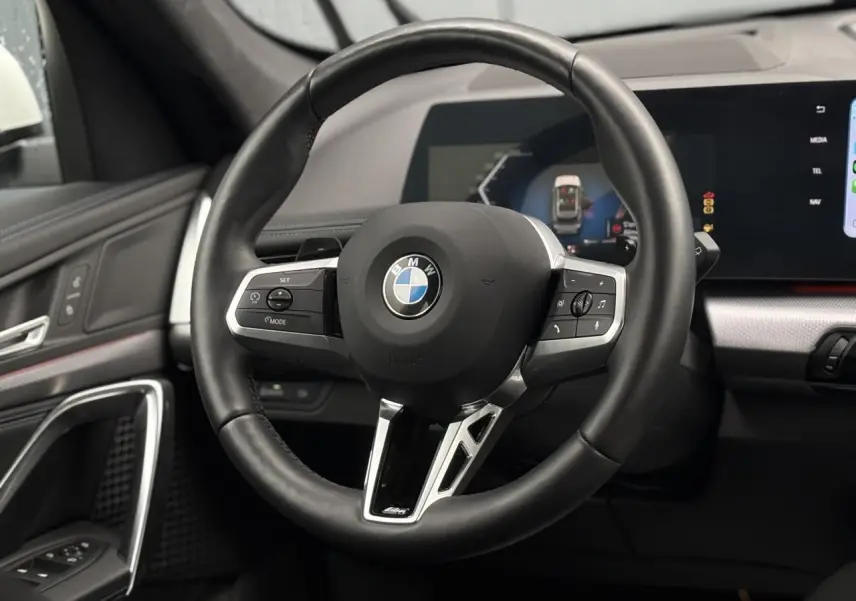 Vue rapprochée du volant cuir noir M avec commandes multifonctions du BMW X1 sDrive 20d blanc Mineral 2025.