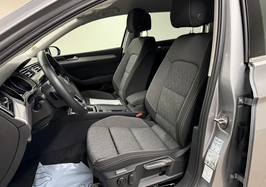 Vue intérieure côté conducteur de la Volkswagen Passat SW 2024 gris pyrite, sièges tissu gris et tableau de bord moderne.