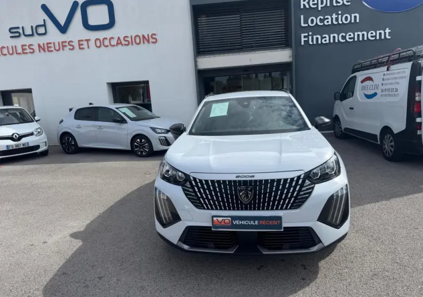Vue avant d’un Peugeot 2008 blanc 2024 avec calandre noire et barres de toit, stationné devant un garage.