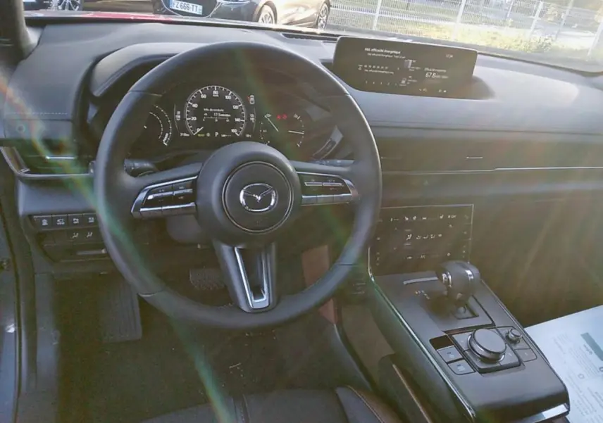 Vue intérieure du tableau de bord et volant du Mazda MX-30 gris, avec écran central et levier de boîte automatique visible.