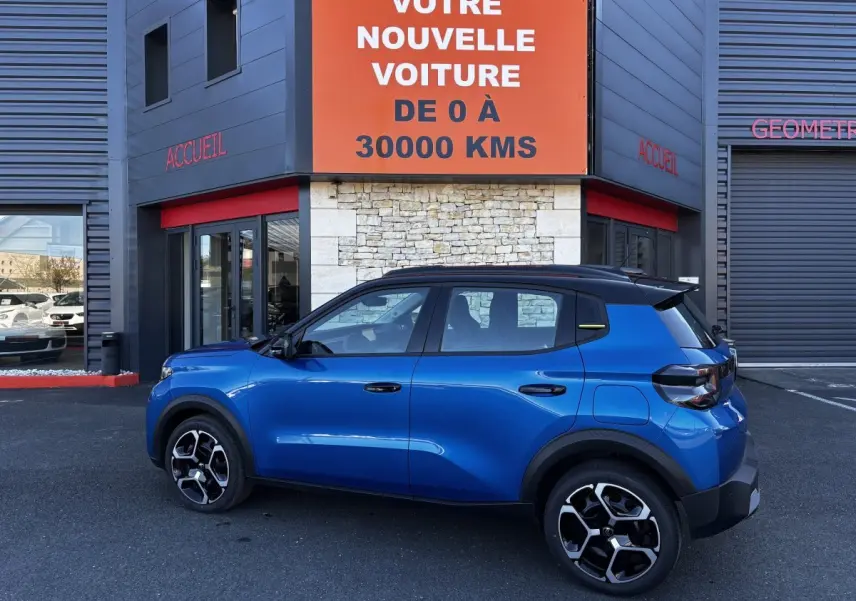 Vue latérale droite d'une Citroën C3 100 BVM PLUS bleu avec toit noir et jantes distinctives, devant un bâtiment commercial.