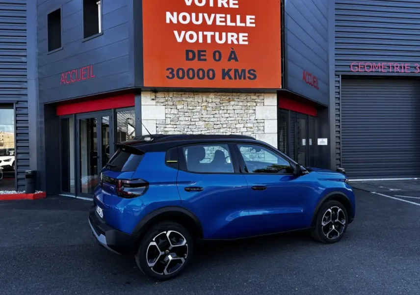 Vue de profil droit d'une Citroën C3 100 BVM PLUS bleu avec toit noir, roues alliage et feux arrière LED, devant un garage.