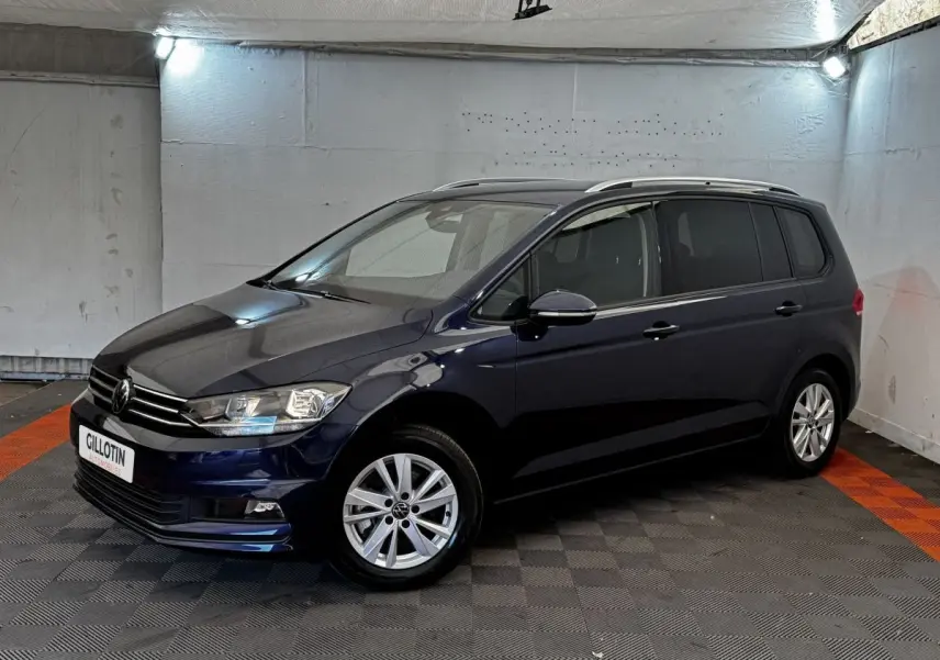 Volkswagen Touran bleu 3/4 avant droit en intérieur, avec jantes alliage et rails de toit chromés visibles.