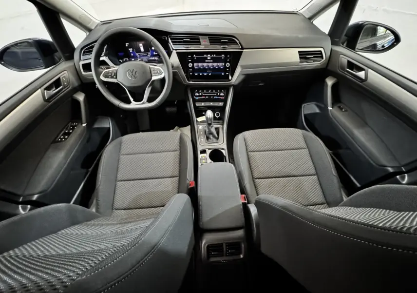 Vue intérieure avant du Volkswagen Touran 2024 avec tableau de bord noir et sièges tissu gris foncé.